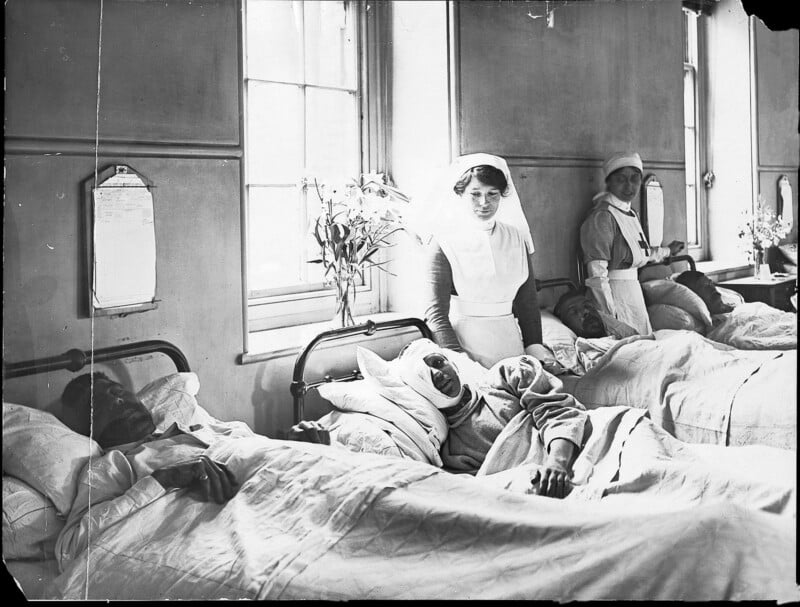 Black-and-white photo of three nurses attending to four patients in hospital beds by a sunlit window; the patients are covered with blankets and one has a bandaged face. Flowers sit on the windowsill.
