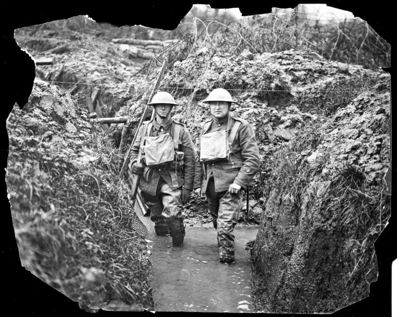 Two soldiers wearing helmets and gear stand knee-deep in water inside a muddy World War I trench, surrounded by rough earth walls and barbed wire.