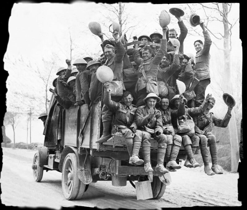 A group of smiling World War I soldiers sit atop and hang off a crowded military truck, waving their hats joyfully as they travel down a rural dirt road lined with bare trees.