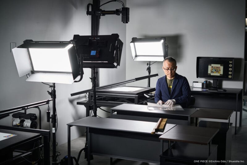 Un hombre con gafas y guantes se sienta en una mesa en un estudio con luces brillantes y equipo de cámara. El monitor muestra una imagen en color. El espacio de trabajo parece organizado y profesional.