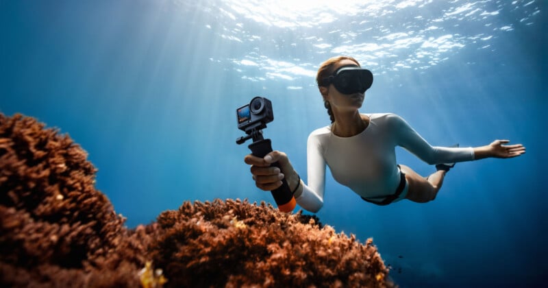 A person wearing a white rash guard and snorkel mask swims underwater, holding an action camera near coral reefs, with sunlight streaming through the water above.