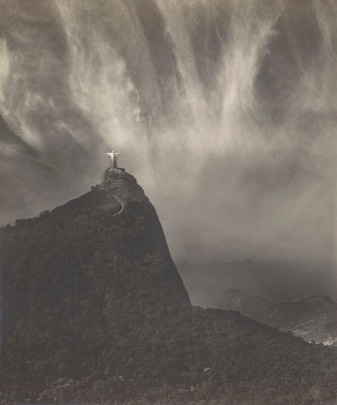 A black and white photo of the Christ the Redeemer statue atop Mount Corcovado in Rio de Janeiro, surrounded by lush forest and dramatic, wispy clouds in the sky above.