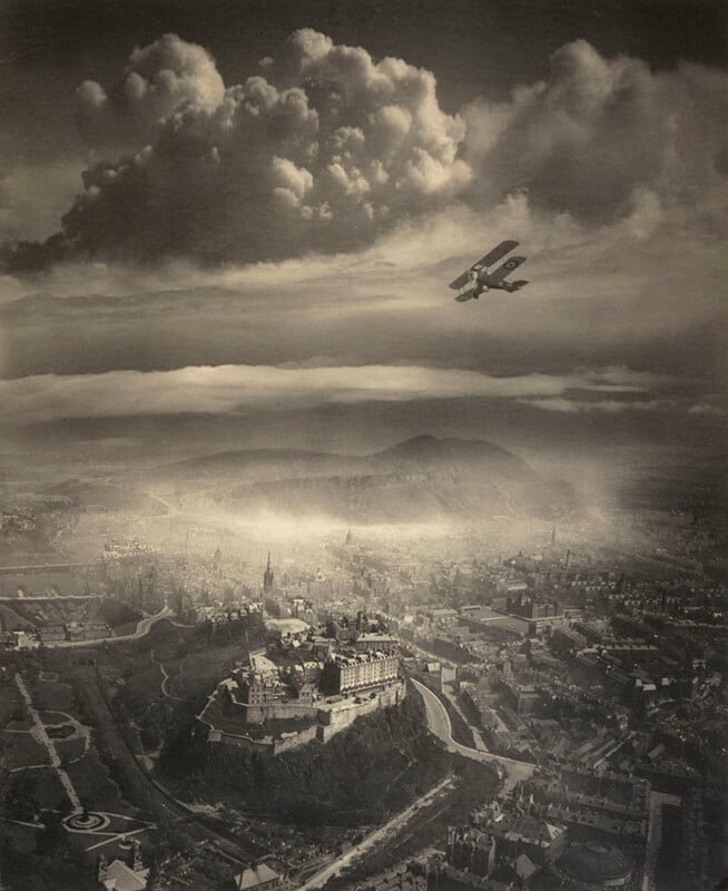 A vintage, sepia-toned aerial photograph shows a biplane flying above a sprawling city with a prominent castle or fortress on a hill, dramatic clouds overhead, and distant mountains on the horizon.
