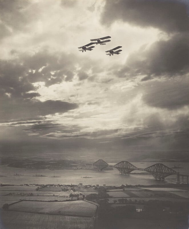 Four vintage biplanes fly in formation over a river, with a large iron bridge spanning the water below and dramatic clouds filling the sky. Fields, houses, and distant land are visible beneath the aircraft.