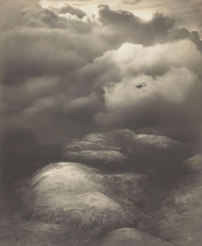A vintage biplane flies above snow-covered hills, with dramatic clouds dominating the sky and sunlight streaming through, casting shadows on the landscape below.