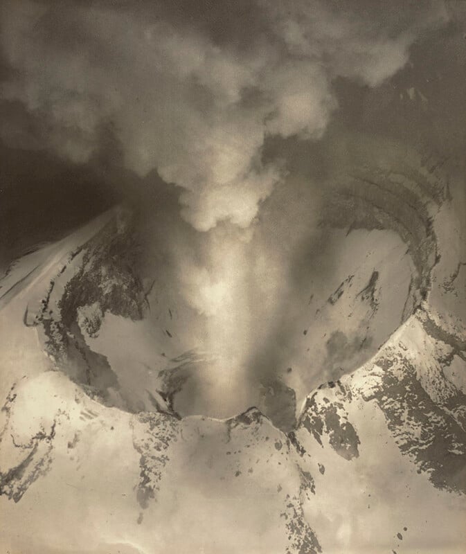 Aerial view of a volcanic crater with smoke and ash billowing upward, surrounded by snow-covered slopes and rugged terrain. The image appears in sepia tones, giving it a vintage look.