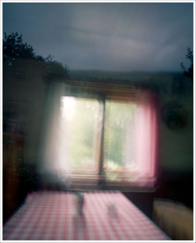 A blurry image of a window with soft light streaming in, behind a table covered with a red and white checkered tablecloth. The scene has an ethereal, dreamy quality with indistinct objects on the table.