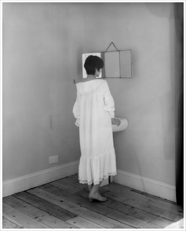 A person in a long, white nightgown stands barefoot on a wooden floor, facing a small wall-mounted mirror above a sink in a plain room with bare walls.