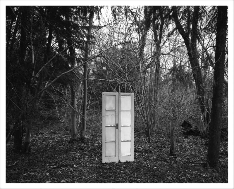 A pair of white double doors stands alone in a leaf-covered, barren forest with bare trees, creating a surreal and mysterious scene in black and white.
