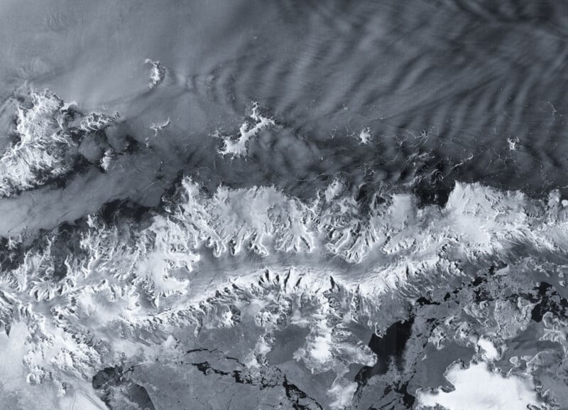 Las imágenes de satélite muestran montañas cubiertas de nieve rodeadas de agua oscura y helada y nubes veteadas. Picos y crestas irregulares dominan el paisaje, creando un marcado contraste con el área circundante.