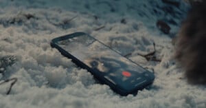 A smartphone lying on snow records a video, showing the face of a furry animal, likely a dog, on its screen. The scene appears outdoors, with twigs and pine needles visible around the phone.