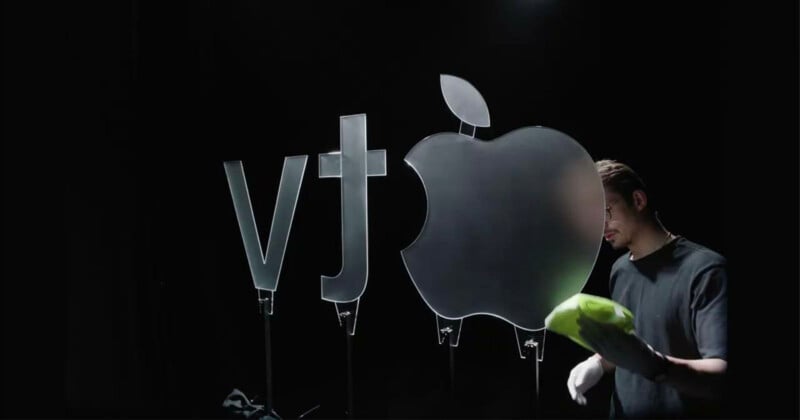 A person wearing gloves cleans a large glass Apple logo next to "vt" symbols, all standing upright against a dark background.