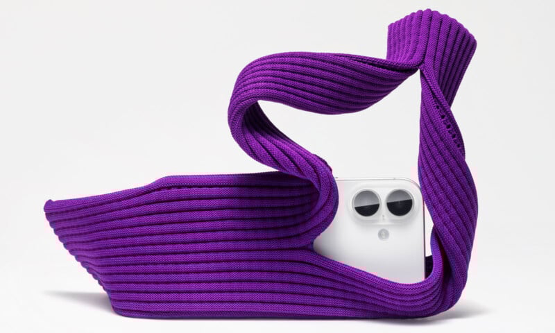 A white smartphone with dual cameras is partially wrapped in and emerging from a sculptural, ribbed, purple fabric against a plain white background.