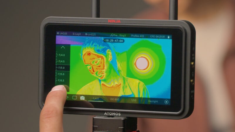 A hand holds a monitor displaying a thermal image of a woman with her head tilted, showing varying heat levels in bright colors, including red, yellow, and green, with a bright circular hotspot behind her.