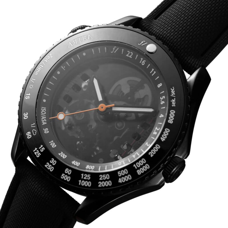 Reloj negro con mecanismo de engranajes visible, marcas y números blancos en el bisel. El reloj cuenta con manecillas de horas y minutos con punta naranja y una correa de tela negra. nombre de marca "Astra" Visible en la parte superior.