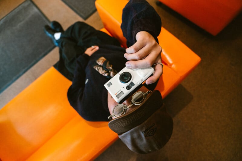 A person wearing a cap and glasses reclines on an orange couch, holding a white camera up toward the viewer, with the image taken from above their head.