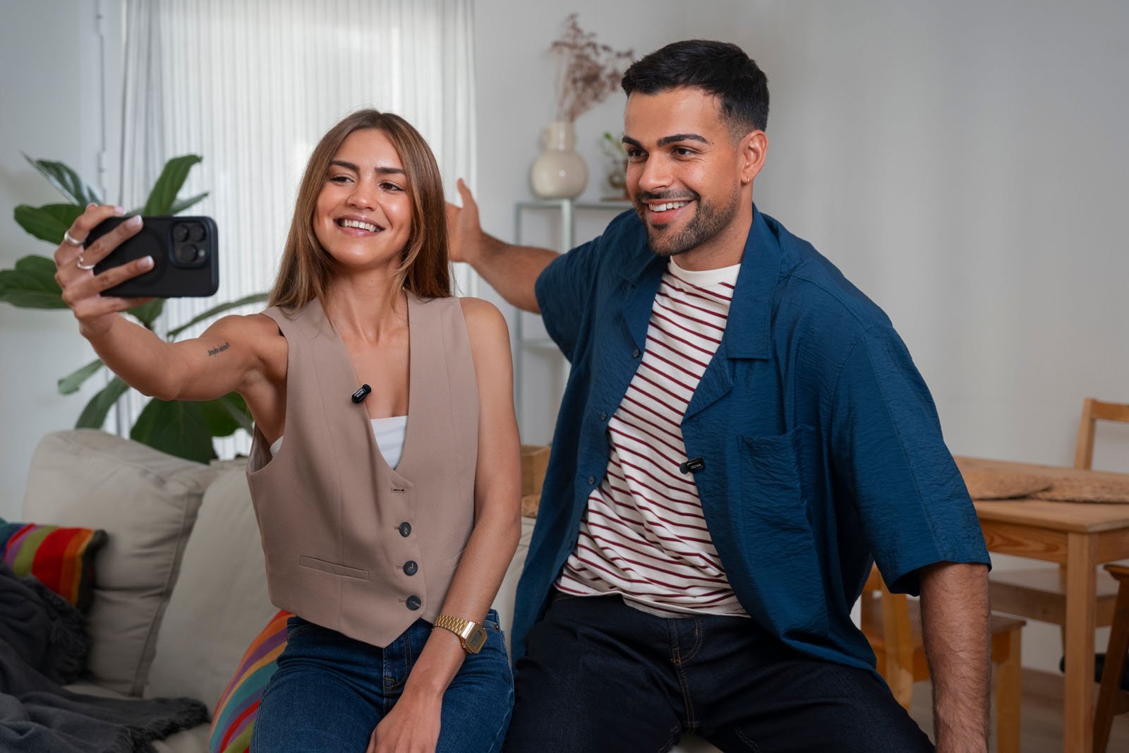 A smiling woman and man sit on a couch in a bright living room, taking a selfie together. The woman holds the phone, and both look happy and relaxed. There are chairs and a table in the background.