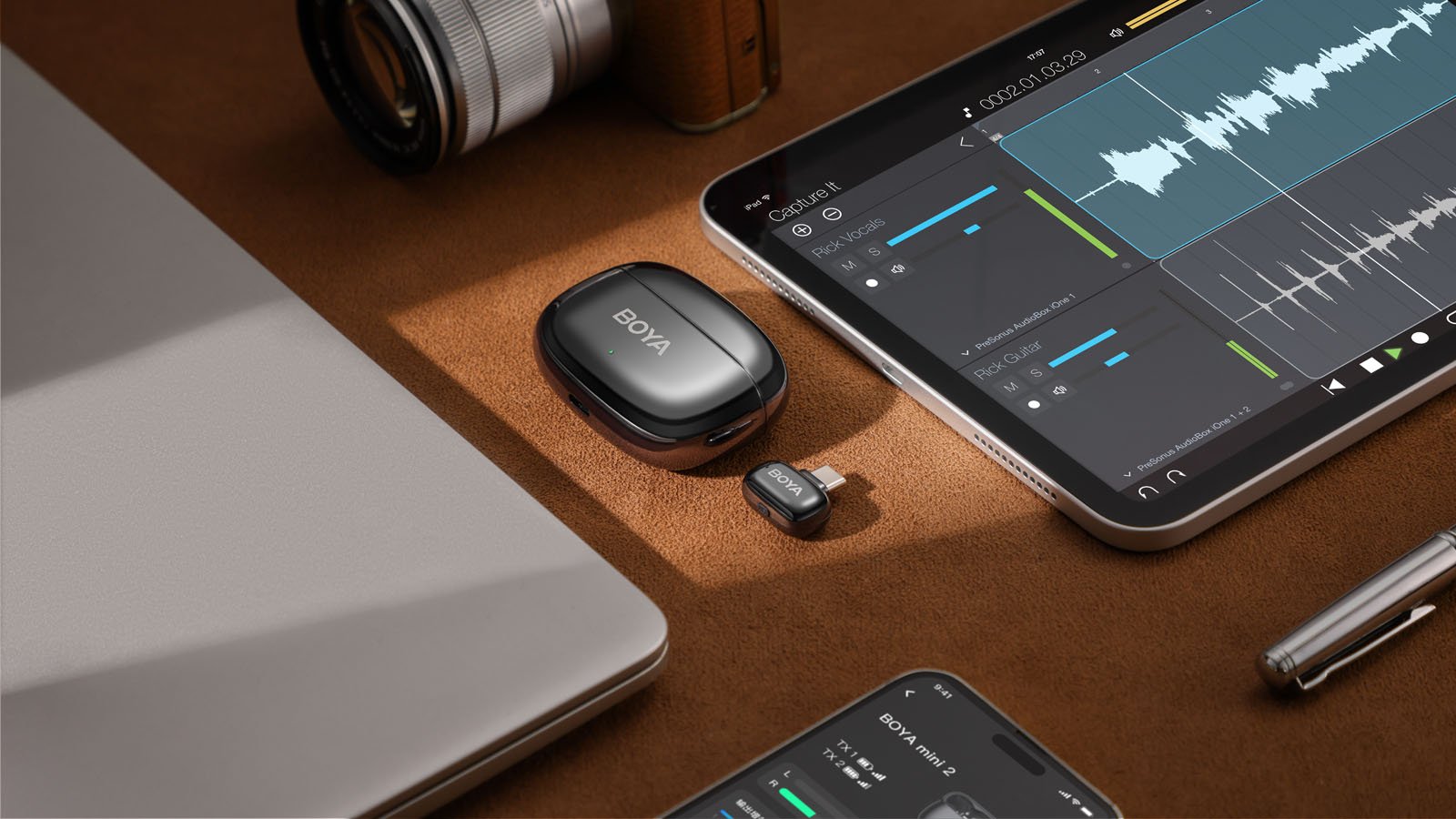 A wireless BOYA microphone set is placed on a brown surface next to a closed laptop, tablet displaying audio editing software, a smartphone, a camera lens, and a pen.