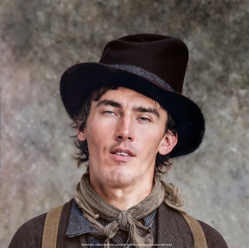 Un joven con un sombrero marrón oscuro, una chaqueta marrón y una bufanda gris se encuentra frente a un fondo neutro texturizado. Tiene piel clara, ojos azules y una expresión relajada.