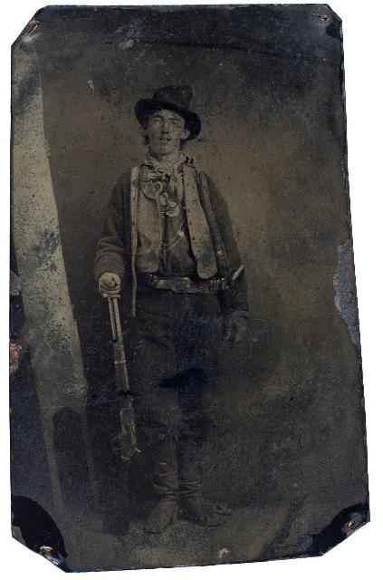 Una fotografía antigua en tonos sepia muestra a un joven vestido con atuendo del viejo oeste, sombrero, pañuelo, chaqueta y botas, de pie contra un fondo liso, sosteniendo un rifle a su lado. Los bordes de la foto están desgastados y oscurecidos.