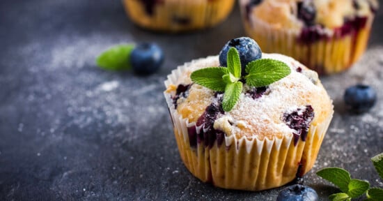 A blueberry muffin topped with powdered sugar, a fresh blueberry, and mint leaves sits on a dark surface, surrounded by scattered blueberries and mint.