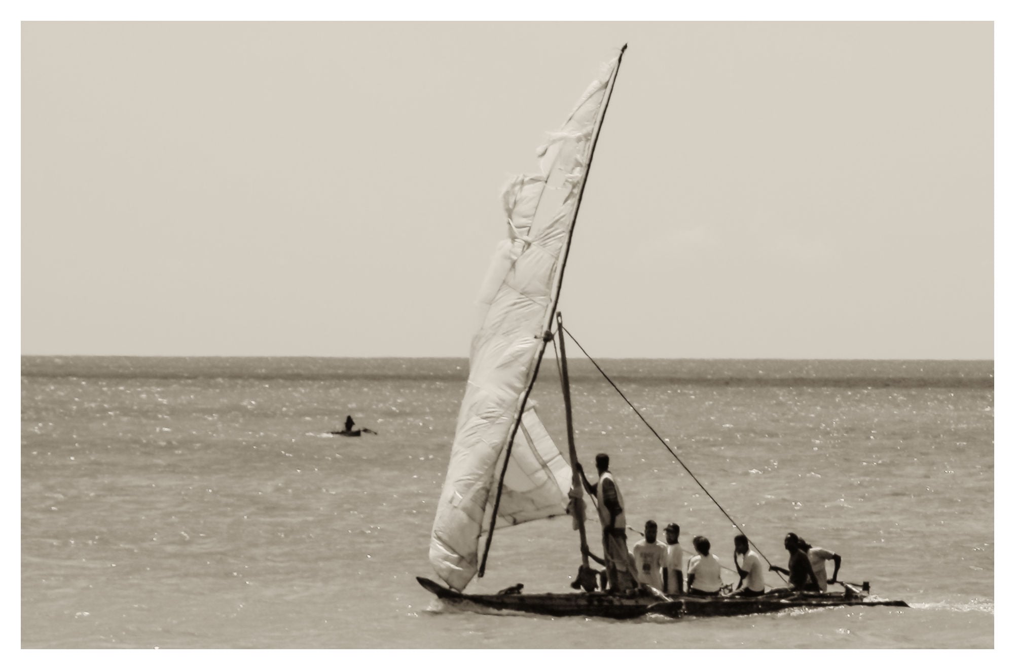 Un grupo de personas navegaba por las tranquilas aguas en un barco de madera con una gran vela blanca. La foto está en blanco y negro y muestra a una persona sentada en un barco más pequeño al fondo.