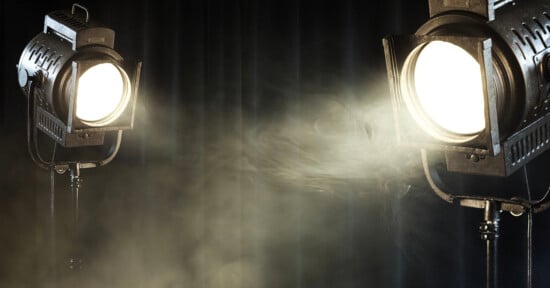 Two large studio spotlights shine bright beams through hazy mist, illuminating the dark, smoky atmosphere against a black curtain backdrop.