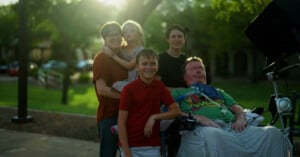 A group of five people, including a child, stand and smile outdoors near a man in a wheelchair equipped with medical equipment. Sunlight filters through the trees in the background, creating a warm and happy atmosphere.