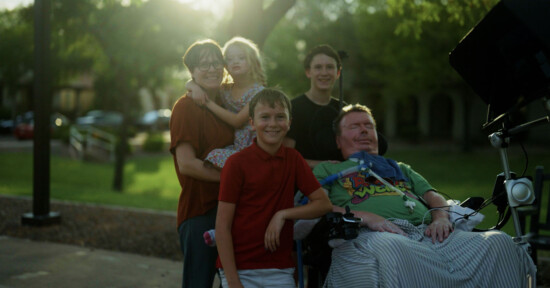 A group of five people, including a child, stand and smile outdoors near a man in a wheelchair equipped with medical equipment. Sunlight filters through the trees in the background, creating a warm and happy atmosphere.