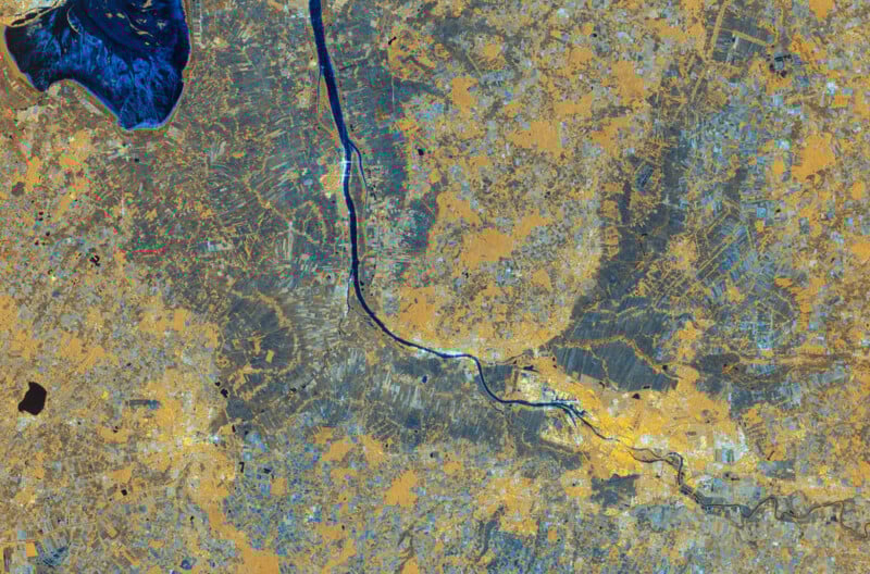 La imagen de satélite muestra un río serpenteando a través de un paisaje con fuentes de agua azules, áreas color canela y gris que representan la tierra y patrones visibles de campos, caminos y desarrollo urbano.