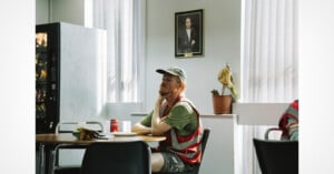 A man in a reflective vest sits at a table with snacks and a can of Coke, appearing thoughtful. Sunlight filters through vertical blinds. A vending machine, potted plant, and a framed portrait are in the background.