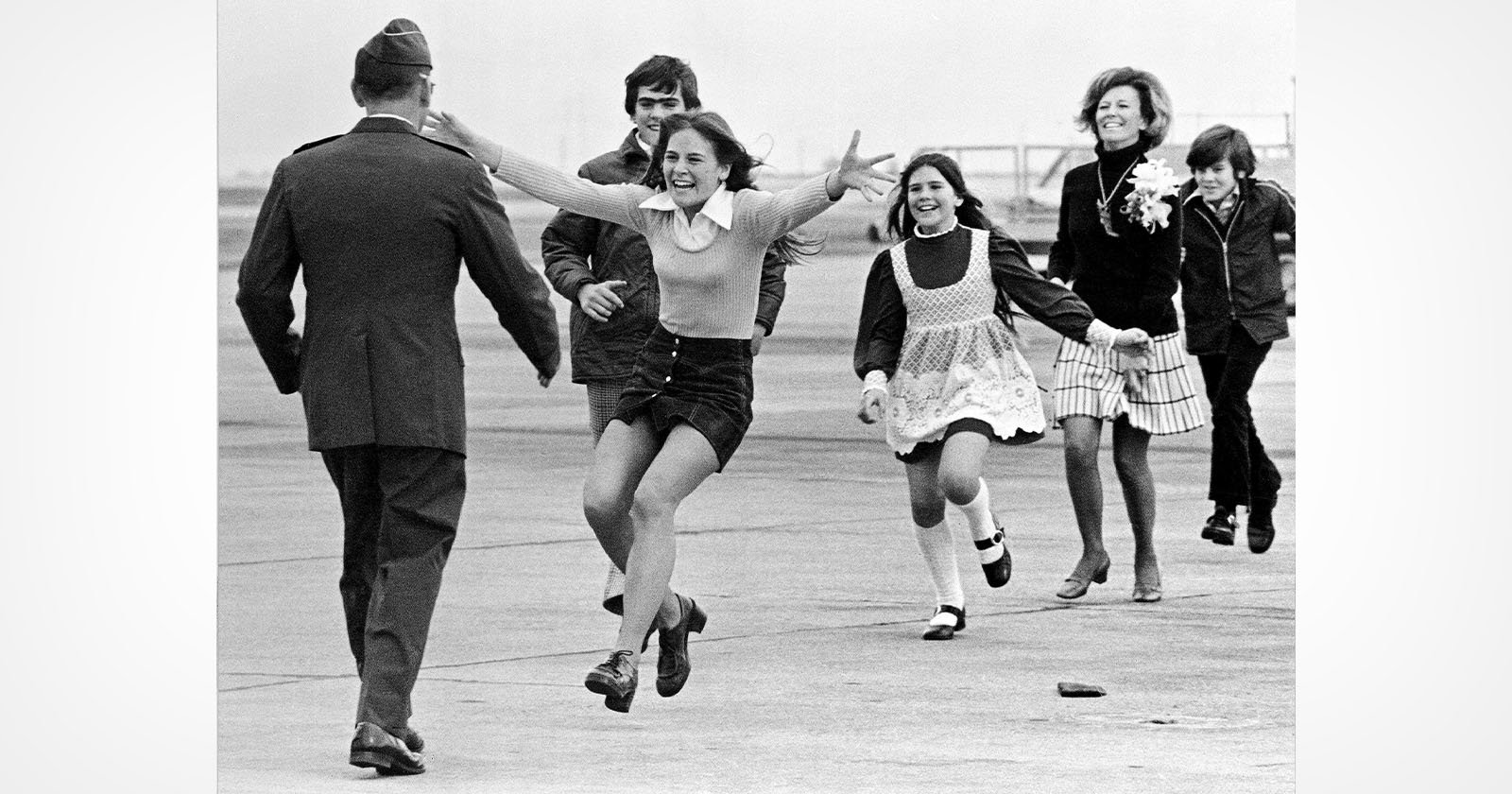 A young girl runs with open arms toward a uniformed man, likely her father, at an airport while other smiling family members follow joyfully behind her.