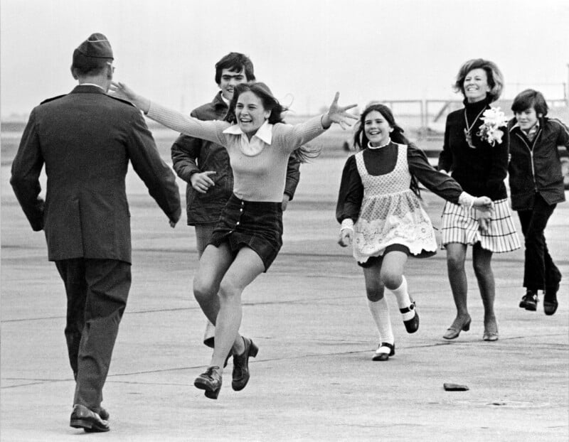 En la pista del aeropuerto, un hombre con uniforme militar camina hacia una niña feliz que corre con los brazos extendidos, seguida por su familia sonriente.