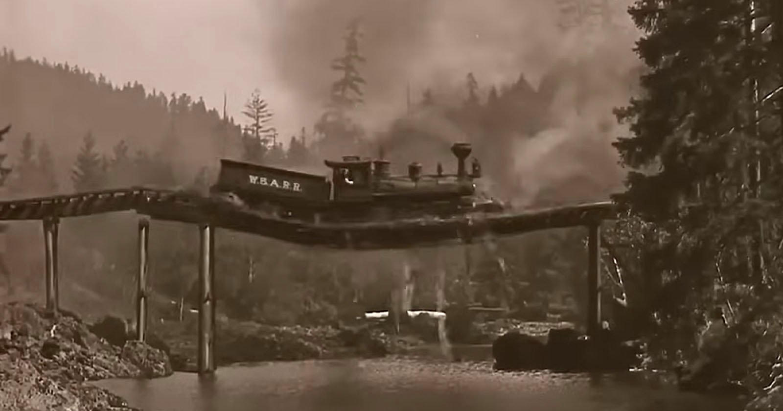 A vintage steam train crosses a precarious, warped wooden bridge over a river, with smoke billowing and tall trees and hills in the background. The scene is in black and white.