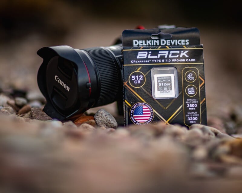 A Canon camera with a lens cap sits on rocky ground next to a Delkin Devices BLACK 512GB CFexpress memory card box. The image is focused on the memory card packaging, with blurred rocks in the background.