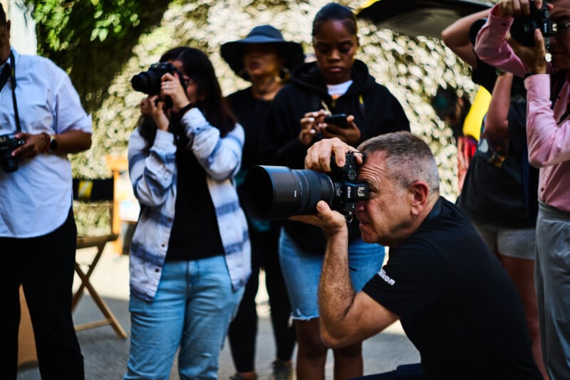 A group of people, some standing and one kneeling, focus intently as they take photographs with cameras outdoors. The background is blurred, and sunlight filters through trees above.