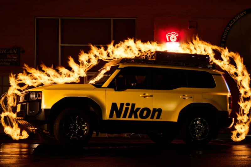 A yellow SUV with a large “Nikon” logo is parked at night. Flames arc around the vehicle, creating a dramatic effect. A glowing red camera sign is visible on the roof. The background is dimly lit.