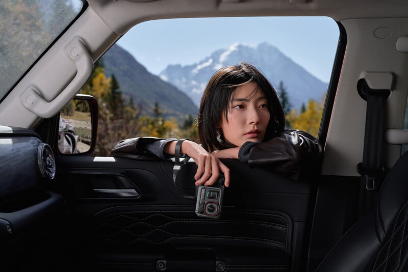 Una mujer de pelo corto y negro se asomó por la ventanilla del coche sosteniendo una cámara. Bajo un cielo despejado, las montañas nevadas y los árboles del fondo son claramente visibles.
