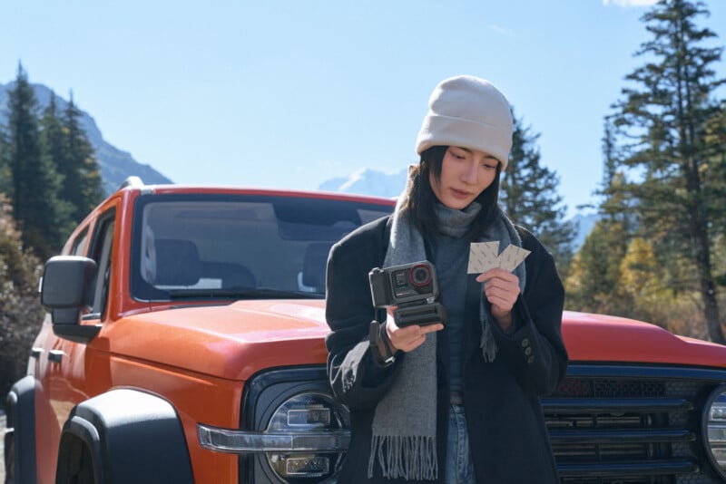 Una mujer con gorro y bufanda se paró frente a un todoterreno naranja, sosteniendo una cámara y mirando varias fotografías instantáneas, con árboles y montañas de fondo.