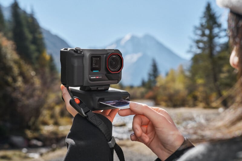 Un hombre que sostiene una pequeña cámara está imprimiendo una fotografía con un pintoresco paisaje montañoso y árboles al fondo en un día soleado.
