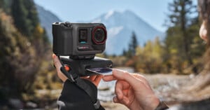 A person inserts a memory card into an action camera mounted on a tripod outdoors, with a blurred background of trees, a river, and snow-capped mountains.