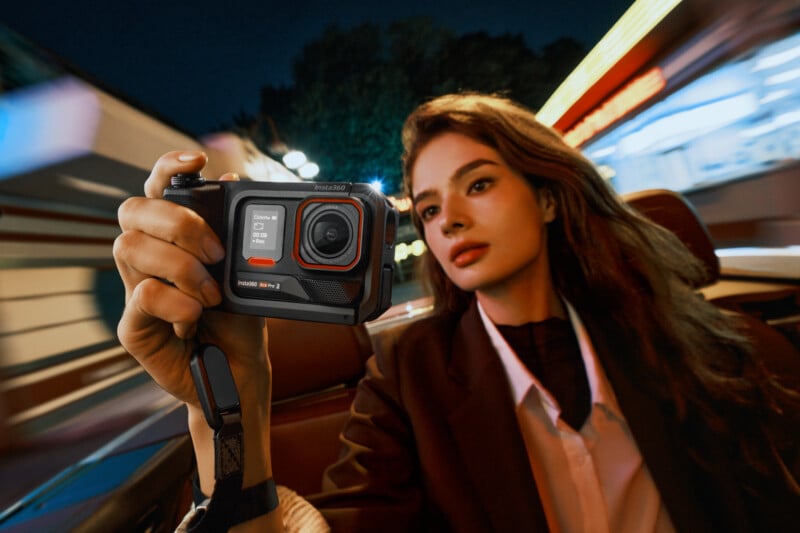 Una mujer de pelo largo sentada en un automóvil en movimiento por la noche, sosteniendo una pequeña cámara de acción frente a su rostro para capturar las luces vibrantes y borrosas de la ciudad y el movimiento circundante.