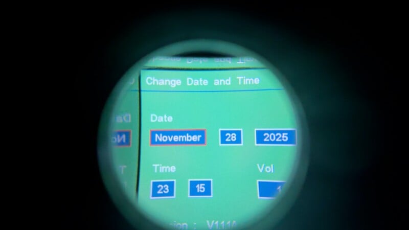 Una pantalla digital vista a través de una lente circular muestra la configuración de fecha y hora: 28 de noviembre de 2025, con opciones de hora que muestran 23 y 15. La interfaz tiene un fondo verde y botones azules.