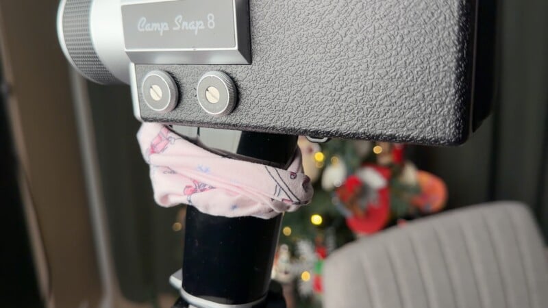Primer plano de una cámara Camp Snap 8 negra montada en un trípode con una tela estampada rosa envuelta alrededor. Al fondo se puede ver un árbol de Navidad vagamente decorado con luces y adornos.