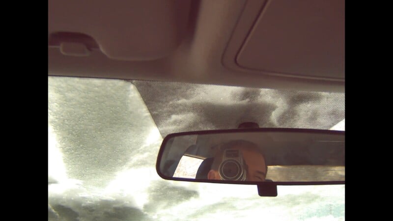 Mirando el cielo nublado desde el interior del coche a través del parabrisas, el espejo retrovisor refleja a un hombre sosteniendo una cámara para tomar fotografías.