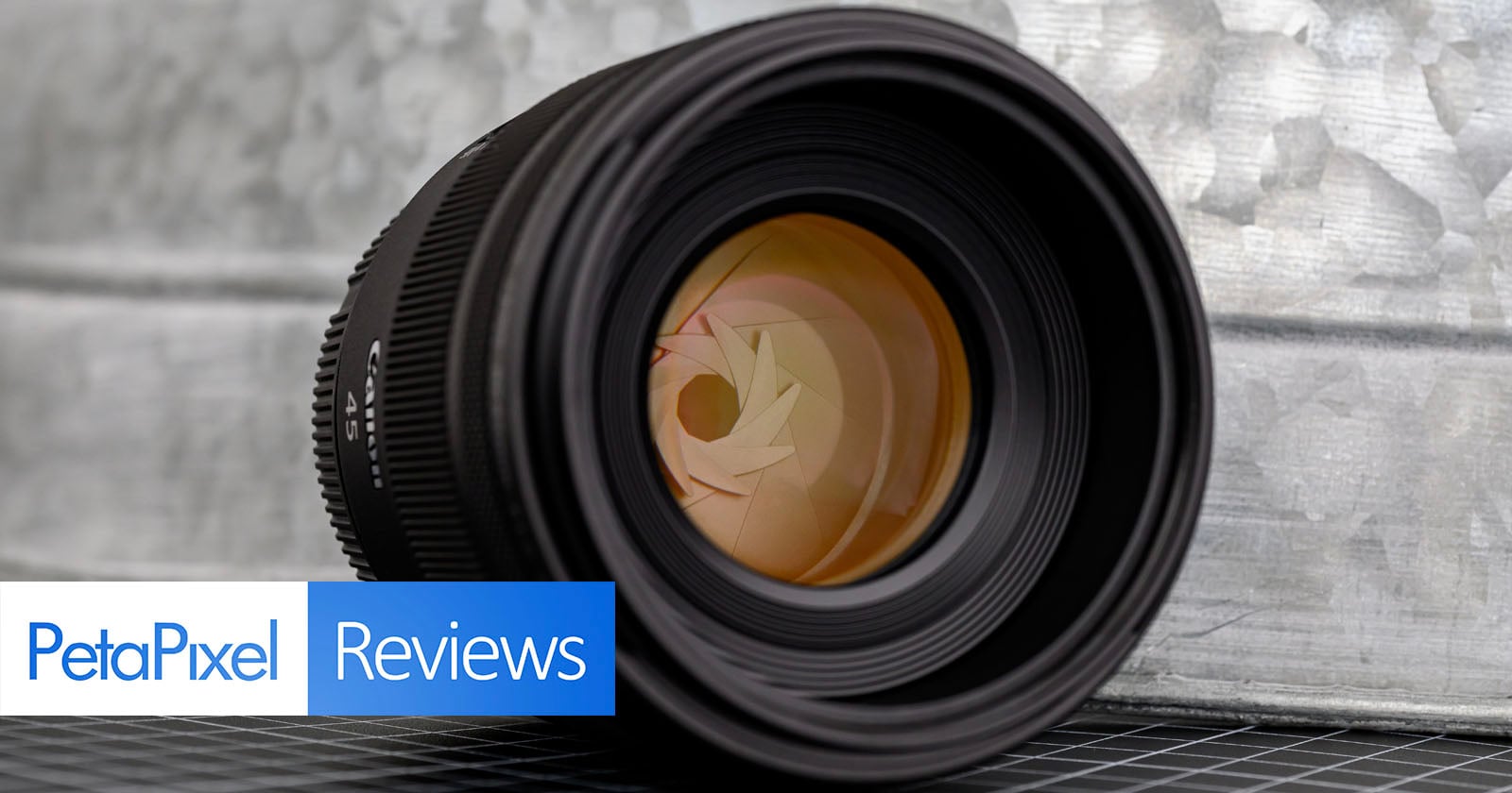 A close-up of a camera lens positioned on its side, with the aperture blades visible. The background features a textured metallic surface and a grid-patterned surface beneath the lens. "PetaPixel Reviews" is overlaid on the bottom left.