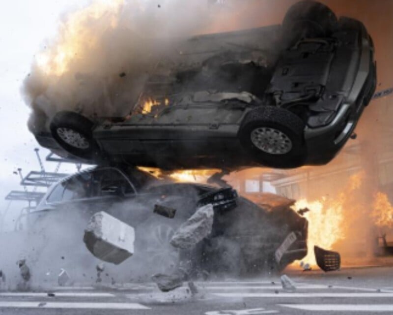 Two cars collide violently at an intersection, with one car flipping upside down in mid-air and debris flying. Flames and smoke erupt in the background, indicating a severe explosion.