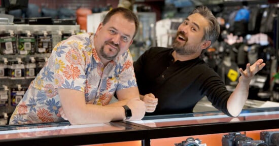 Two men stand behind a glass display counter in a camera store; one wears a floral shirt and leans forward, the other in a black shirt gestures playfully with a smile. Camera gear and accessories are visible in the background.