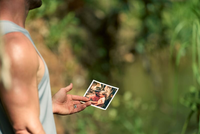 Un hombre que lleva una camisa sin mangas sostiene una fotografía al aire libre. En la foto, un hombre y un niño están sentados juntos. El fondo es verde y borroso, lo que sugiere un entorno natural como un bosque o un parque.