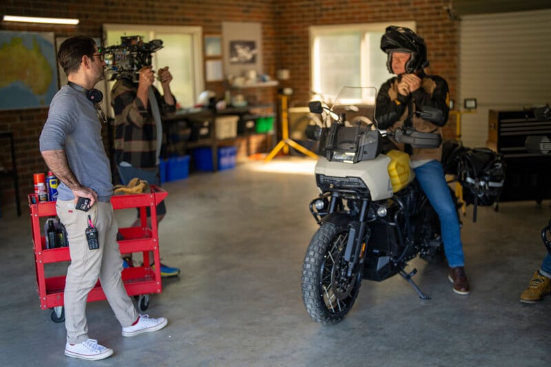Un hombre está sentado en una motocicleta en el garaje, ajustándose el casco. Otro hombre estaba cerca con un walkie-talkie mientras un tercer hombre filmaba la escena con una cámara. Al fondo se pueden ver varias herramientas y equipos.